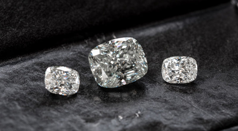 Three loose cushion-cut diamonds in varying sizes arranged on a dark textured surface.