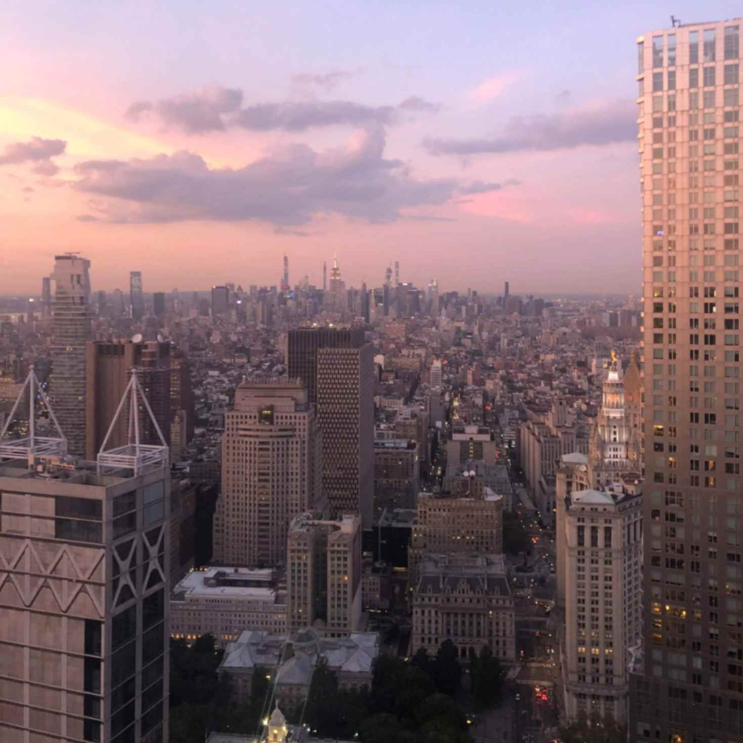 Sunset view of Manhattan with pastel pink skies, tall skyscrapers, and city lights beginning to glow across the skyline.