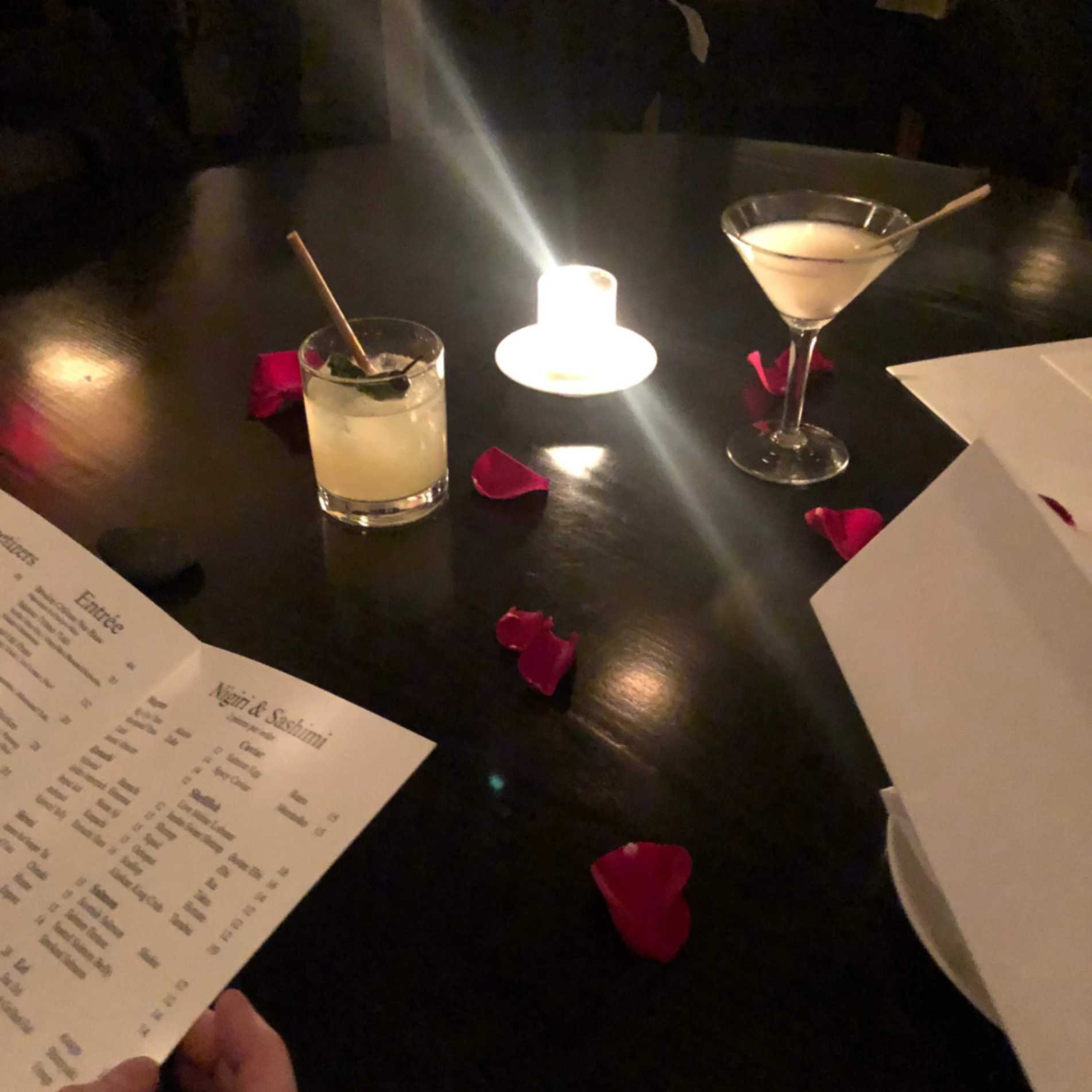 Romantic candlelit dinner table with cocktails, rose petals, and menus, creating an intimate date-night atmosphere