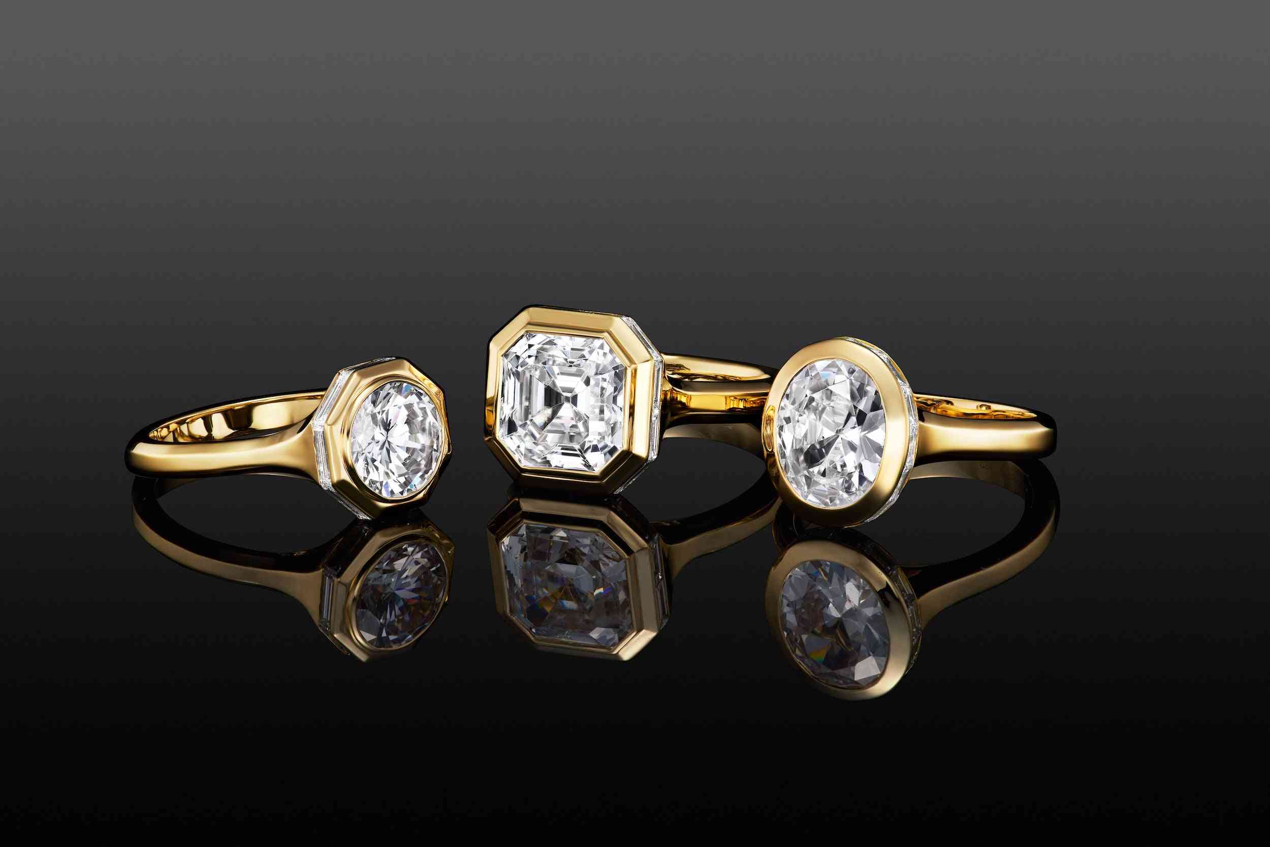 Three yellow gold engagement rings with bezel-set round, Asscher, and oval diamonds against a black reflective background.