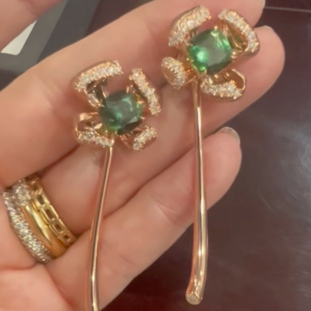 Gold earrings with green gemstones held in a hand against a dark background