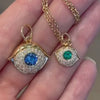 A hand holding two gold evil eye pendants with sapphire and emerald centers.