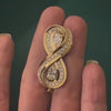 A hand holding a yellow gold infinity brooch with pavé and pear-shaped diamonds.
