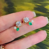 A hand holding gold diamond stud earrings with emerald and diamond ear jackets.