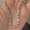 A hand holding a gold tennis necklace with alternating round and emerald cut diamonds.
