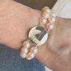 Close-up of woman’s wrist wearing a double-strand pearl bracelet with a round gold fern-engraved locket.