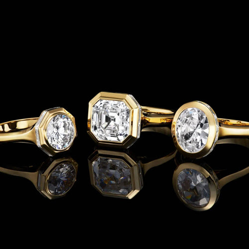 Set of three yellow gold rings featuring oval, asscher, and round cut diamonds displayed on a glossy black reflective surface.